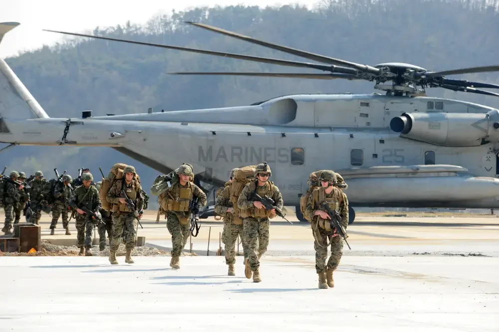 U.S. Marines carrying gear and rifles walking away from a CH-53 helicopter marked 'Marines' during a military exercise