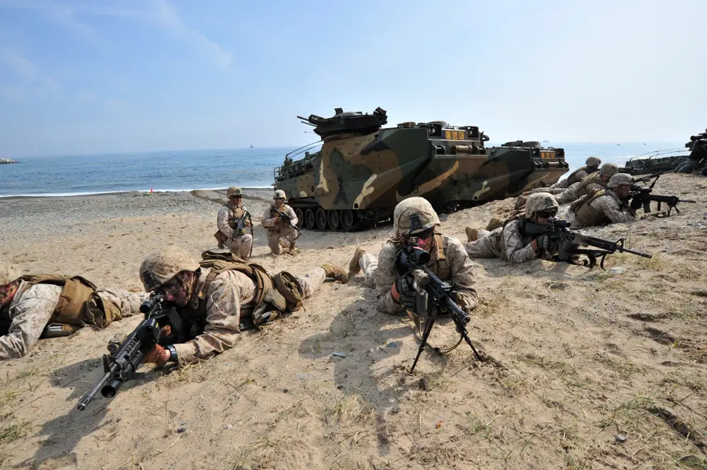 Navy SEALs in tactical gear during a BUD/S beach training exercise, lying prone with rifles near amphibious vehicles.