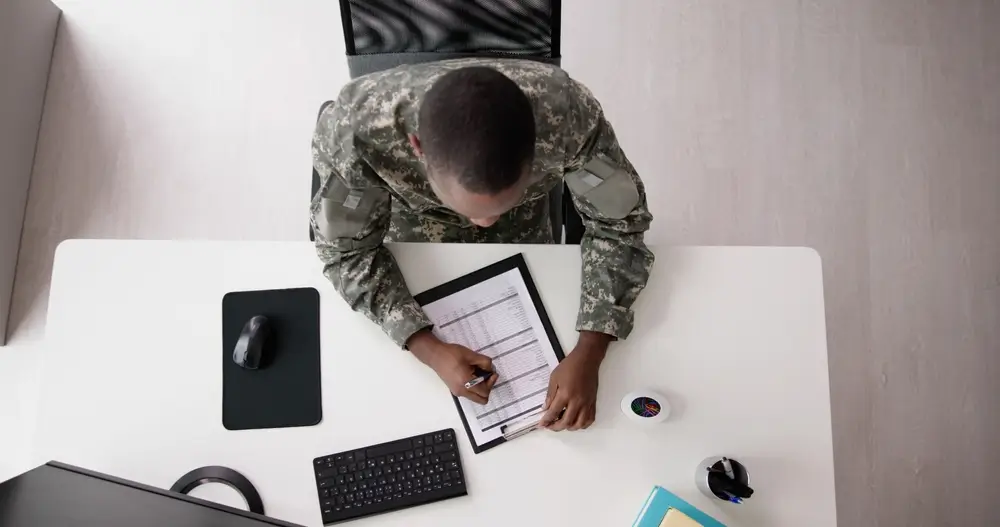 Military applicant in uniform writing on a clipboard at a computer desk in a MEPS computerized exam room.