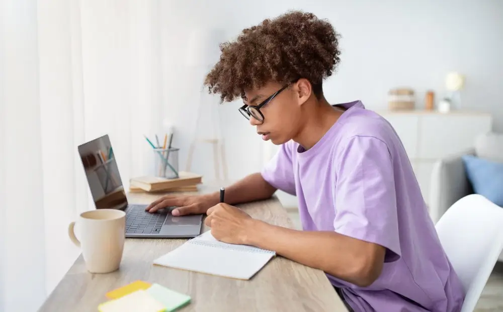 A young man takes an online computer exam on a laptop while writing in a notebook at a home desk.