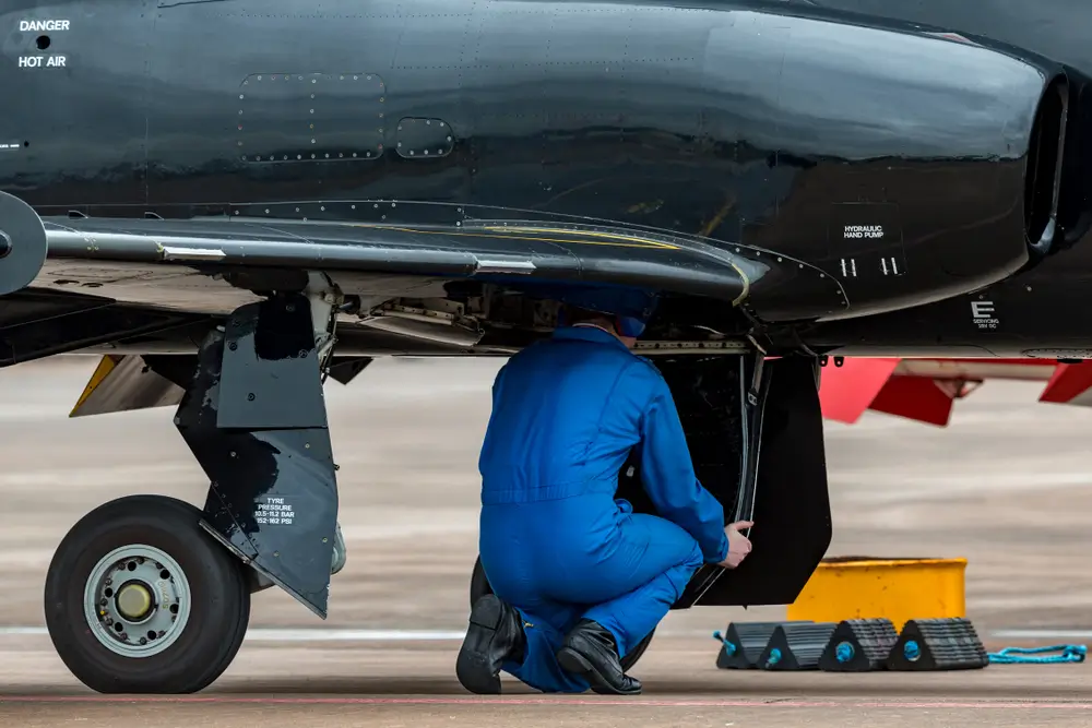 Aviation technician in blue coveralls inspecting the fuselage and mechanical systems of a black military aircraft.