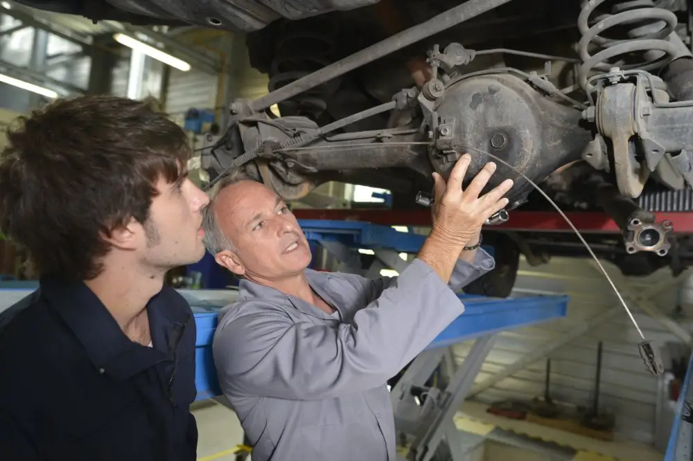 Auto mechanic teaching engine and vehicle mechanics to a student while examining a car's undercarriage on a lift.