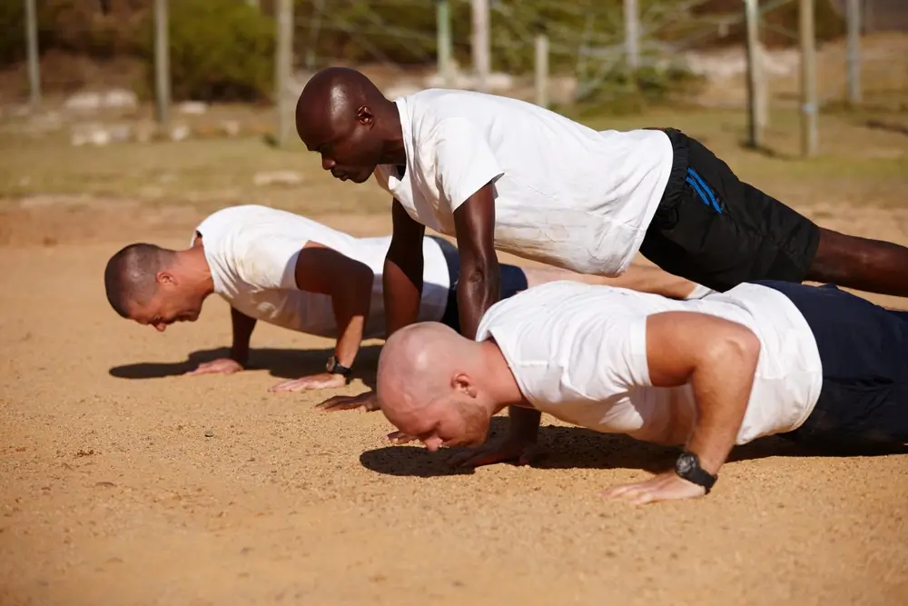 Military recruits in white t-shirts perform push-ups during physical training on an outdoor sandy course.