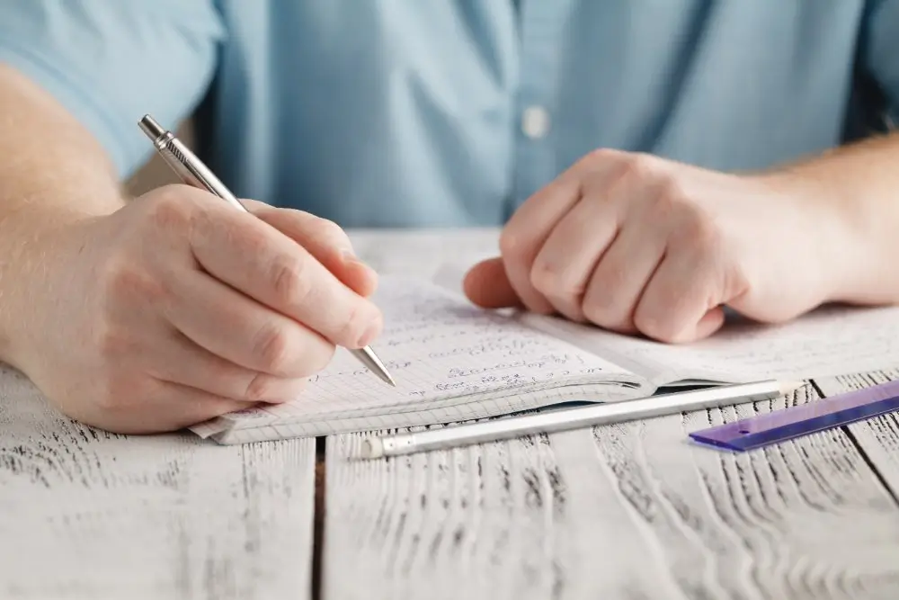 Student writing algebra equations in a notebook at a desk with a pen and ruler during a math exam.